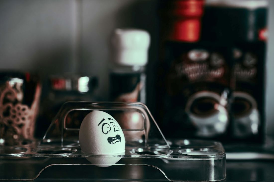 A photograph of single egg with an illustrated face on it. The face is turned to the right and looks apprehensive. The items in the background are out of focus, so it's hard to tell what they are.