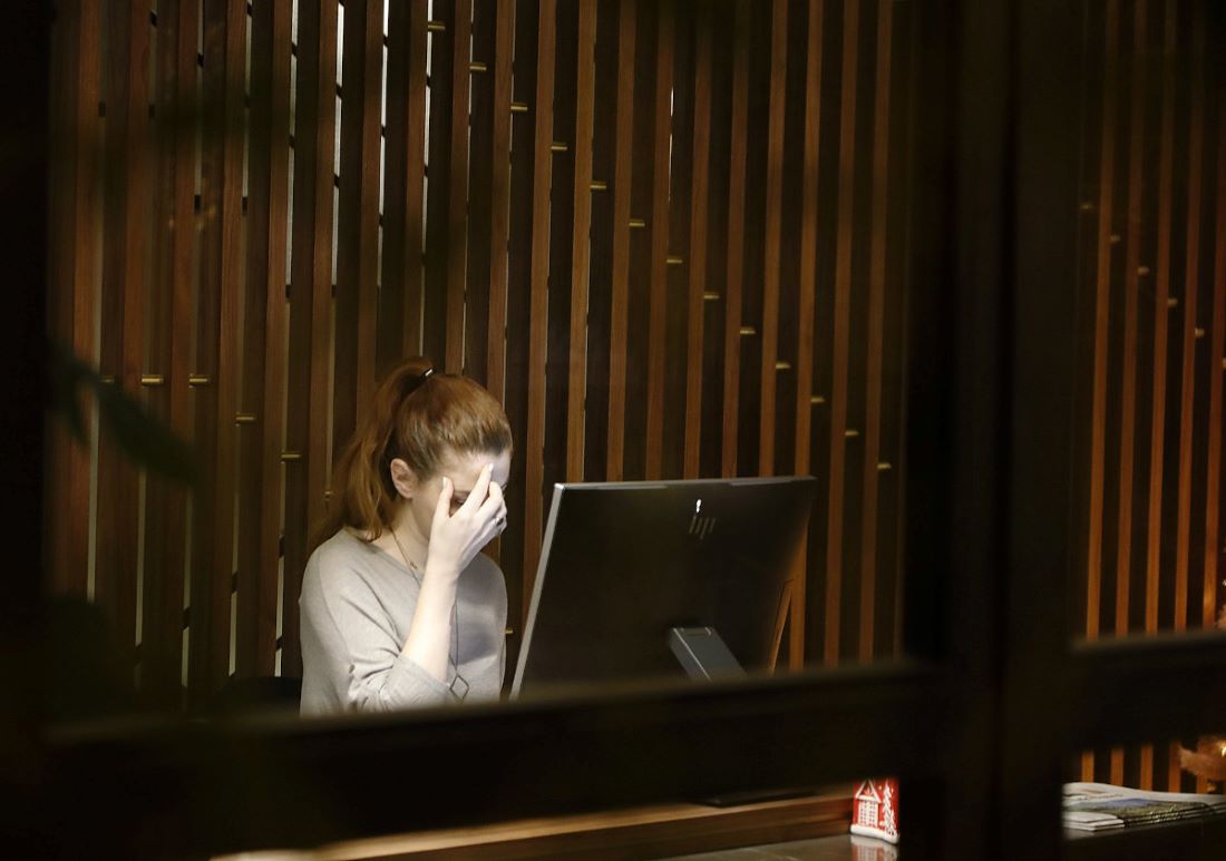 View through a window of a woman sitting at a desk, in front of a computer, rubbing her head as if she has a headache.