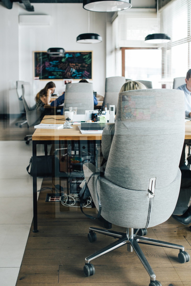 Group of people in a small business, seated in comfortable grey office chairs working at long wooden-topped tables.