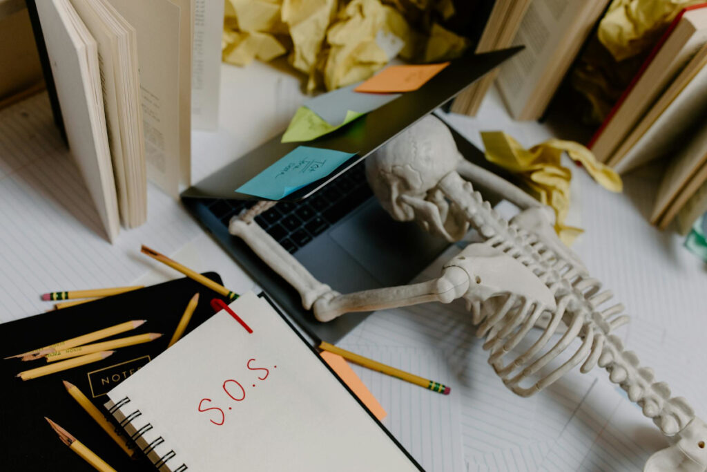 Skeleton face-down on a laptop, surrounded by crumpled paper. "SOS" is written in a notebook lying next to the skeleton.