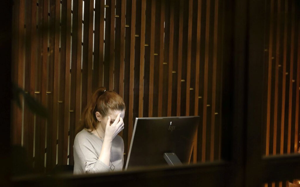 A young woman sits at her desk, rubbing her forehead, against a backdrop of slatted wood.